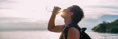 Bir adam gün batımında sahilde bir şişeden su içer. Genç bir adam deniz kenarındaki plastik bir şişeden su içiyor. Portre.