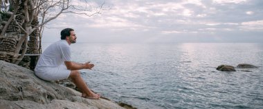 Okyanus kıyısındaki bir taşın üzerinde oturan bir adam. Uçurumun kenarındaki deniz manzaralı beyaz elbiseli genç adam. Adam tropikal bir adada okyanus manzarasını seviyor..