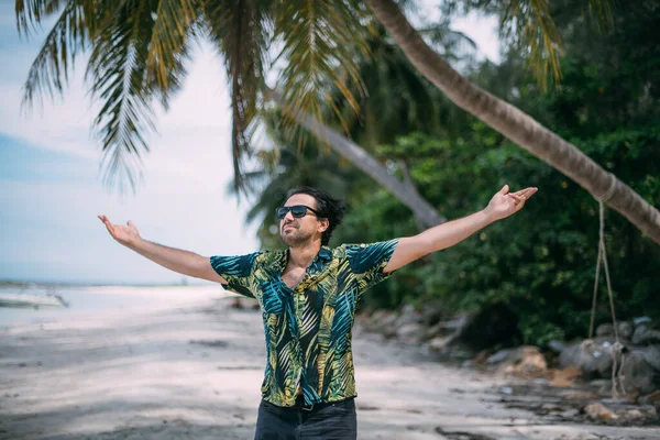 Beautiful man in a Hawaiian shirt walks on a tropical beach. The guy goes near the ocean on the background of palm trees on a tropical island