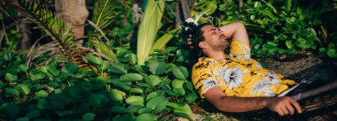 Genç bir adam güneşli bir sahilde bir hamakta yatıyor. Yakışıklı, Hawaii gömlekli bir turist, tropik yeşilliğin arka planında hamakta dinleniyor. Gün batımı ışığı. Tropik ada