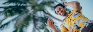 Memnun bir adam elinde telefonla palmiye ağaçlarının arka planında dikiliyor. Tropikal bir adada parlak Hawaii gömlekli genç bir adam. Yakışıklı adam ve parlak olumlu duygular..