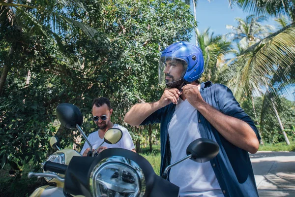 热带岛屿上两个骑摩托车 头戴钢盔的男人 游客们把头盔戴在马路上的棕榈树上 — 图库照片