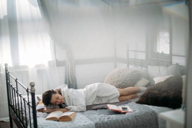 Bornozlu bir kadın elinde kitapla yatakta uyuyor. Genç bir kız dinleniyor, öğleden sonra yatak odasında uyukluyor.