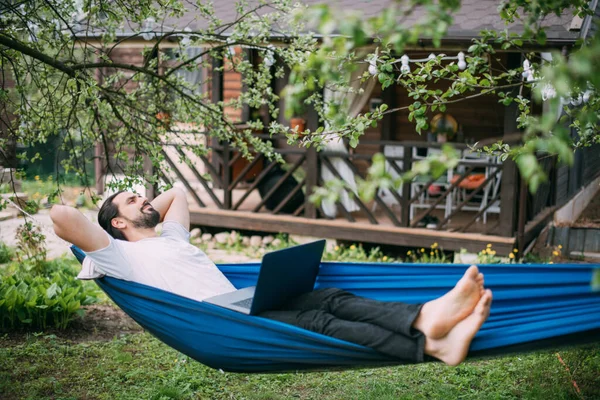 A man works with a laptop in a hammock in a country house. A young guy lies in a hammock with a computer, communicates online, learns remotely under green trees in the country in a summer