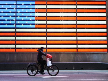 New York, ABD, 24 Mart 2020: Coronavirus salgını sırasında Times Meydanı 'nda Amerika Birleşik Devletleri bayrağı önünde koruyucu maskeli ve bisiklet süren heorik zorunlu işçi