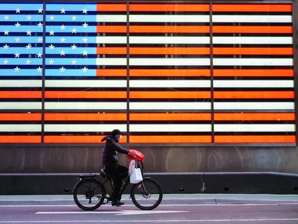 Nova York, Estados Unidos, EUA 24 de março de 2020: trabalhador essencial heórico com máscara protetora andando de bicicleta na frente dos Estados Unidos da América bandeira em tempos quadrados durante o surto de coronavírus Imagens De Bancos De Imagens Sem Royalties