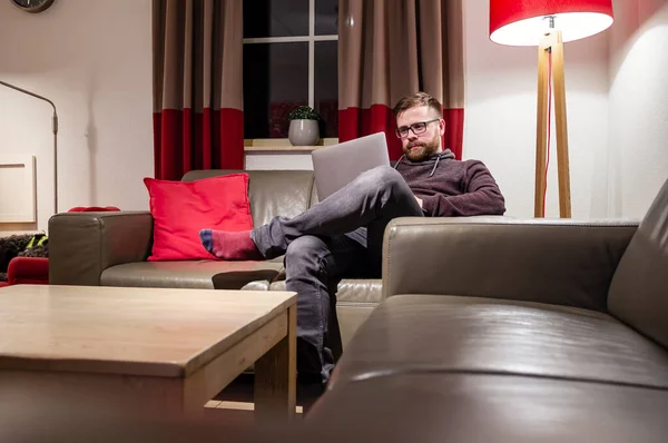 Mannen tittar på sin bärbara dator, läser meddelanden eller arbetar, sitter i en bekväm position på en lädersoffa, i en mysig hemmiljö. — Stockfoto