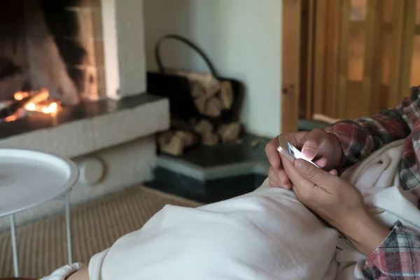 Žena s chytrým telefonem v ruce, sedící na pohovce a přikrytá kostkovanou, na rozmazaném pozadí otevřeného krbu se zapáleným dřevem. — Stock fotografie