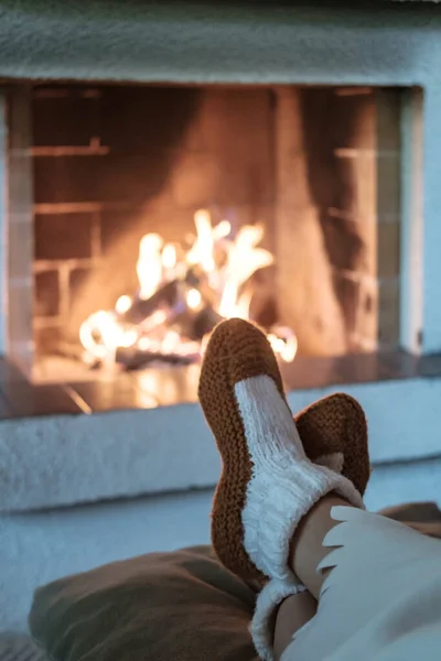 Kvinnliga ben i stickade strumpor täckta med vit filt på suddig bakgrund av öppen spis med tänd ved. Avkoppling, värme och komfort i huset på vinterkvällen. — Stockfoto