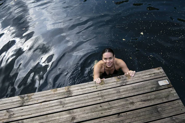 Lächelnde Frau badet in einem klaren See, hält ihre Hände am Rand eines Holzstegs und blickt in die Kamera. — Stockfoto