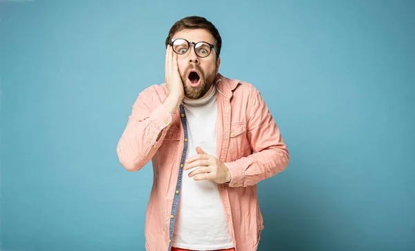 Man in shifted glasses is shocked, he opened mouth, looks at the camera and holds hand to face. Isolated on a blue background. — 스톡 사진