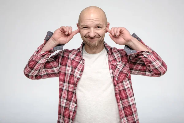 Calvo, hombre sin afeitar cubierto las orejas con los dedos índice para no escuchar, sonríe con astucia y mira a la cámara con los ojos estrechos . — Foto de Stock