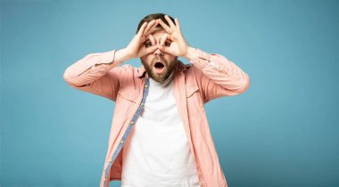 Garip sakallı adam dürbün gibi bir jest yapıyor, elleriyle yüzüne bastırıyor, parmaklarının arasından bakıyor ve şaşırıyor..