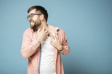 Gözlüklü gergin sakallı adam elleriyle boynu kaşınan cildini kaşındırıyor, acı verici bir şekilde sırıtıyor ve gözlerini kapatıyor. Mavi arkaplan.