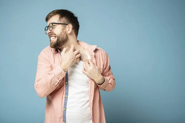 Tense bearded man in glasses scratches itchy skin on neck with his hand, painfully grins teeth and closes eyes. Blue background.