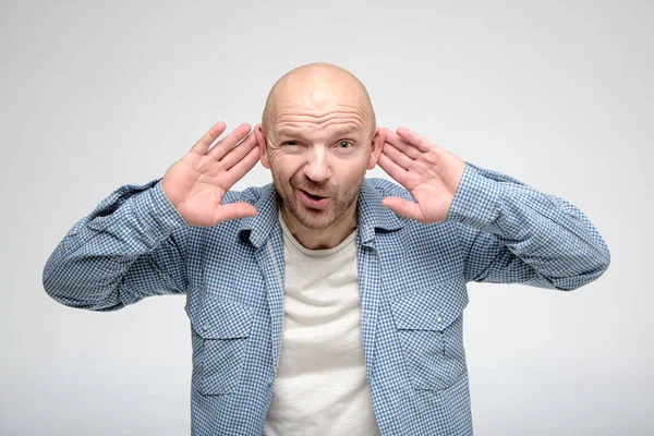 Bald man presses hands to ears and tries to hear something, looking at the camera with a funny expression on face. — Stock Photo, Image