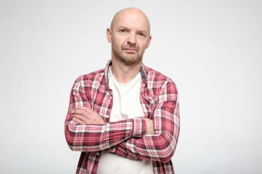 Düşünceli, kaşlarını çatan kel adam kollarını kavuşturup kameraya bakıyor..