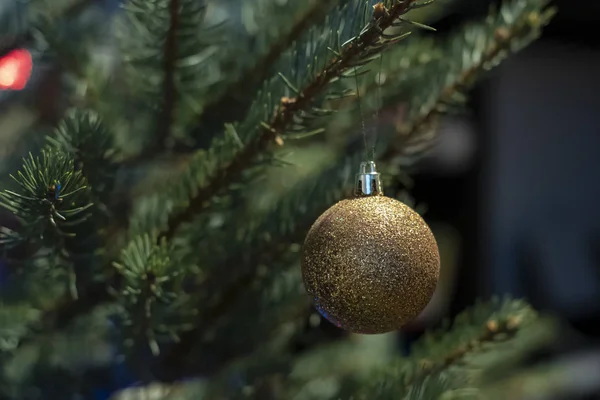 Brilliant gold-colored glass toy ball on a Christmas tree, on a blurred background of garland. — Stock Photo, Image