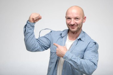 Komik, kel bir adam sanki pazı gibi bir gömlek giyip kameraya bakıyor ve gülümsüyor..