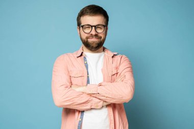 Gözlüklü komik sakallı adam kollarını kavuşturup tuhaf bir şekilde gülümsüyor ve gözlerini kıstırarak kameraya bakıyor..