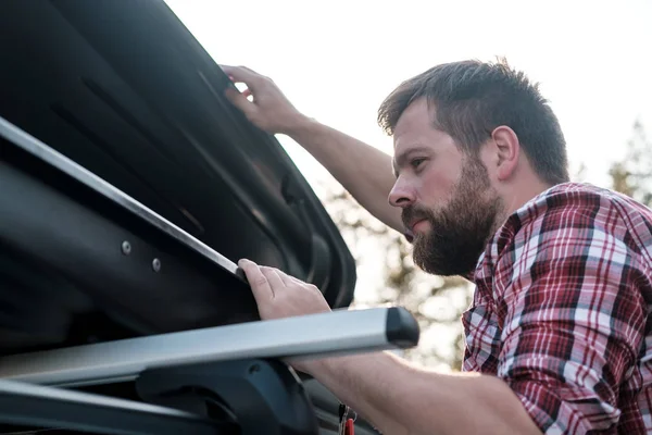 Focused bearded man peers into an open roof rack on a car or in a cargo box. Preparing to leave.