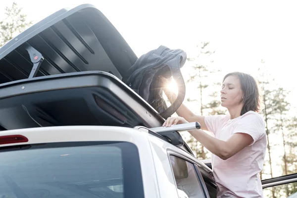 Una mujer seria y enfocada pone cosas en el maletero en el techo de un auto o en una caja de carga. Preparación para unas vacaciones en familia . — Foto de Stock