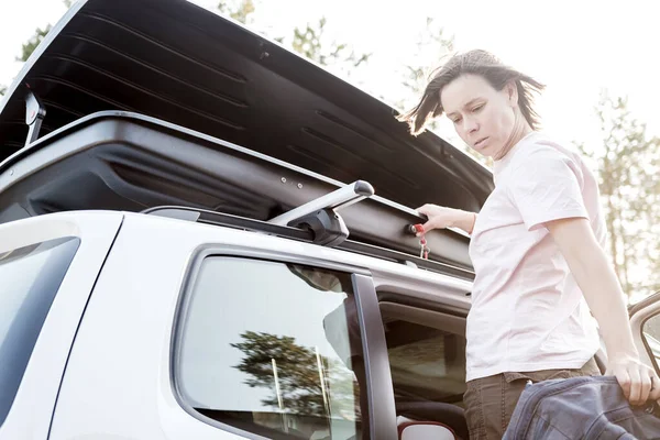 La mujer descarga cosas del maletero o caja de carga, que se encuentra en el techo del coche, en un día ventoso y de verano. Vacaciones familiares . — Foto de Stock