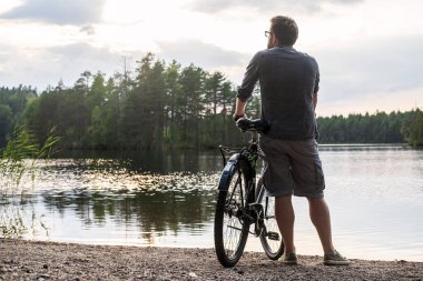 Man with a bicycle stopped on the shore of a forest lake and enjoys the sunset and the beautiful landscape on a summer day.