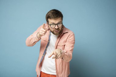 Disgruntled bearded man with glasses accuses and scolds someone, pointing with his index finger and angrily looking at the camera.