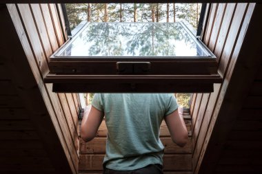 Tavan arasında, açık bir pencereden ağaçların yansıttığı cama yaslanmış bir adamın arkası..