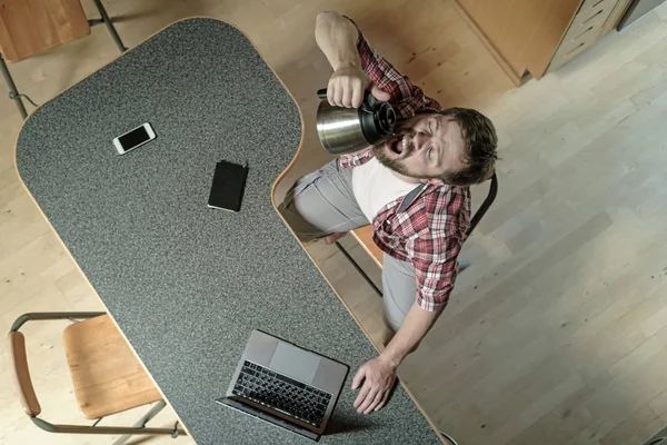 Mann trinkt Kaffee aus einem Wasserkocher, er ist müde und überarbeitet mit einem Laptop, der auf dem Küchentisch liegt. Ansicht von oben. — Stockfoto