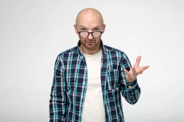 El hombre calvo y sin afeitar mira con inquisición y calma a la cámara sobre las gafas y hace un gesto de mano . — Foto de Stock