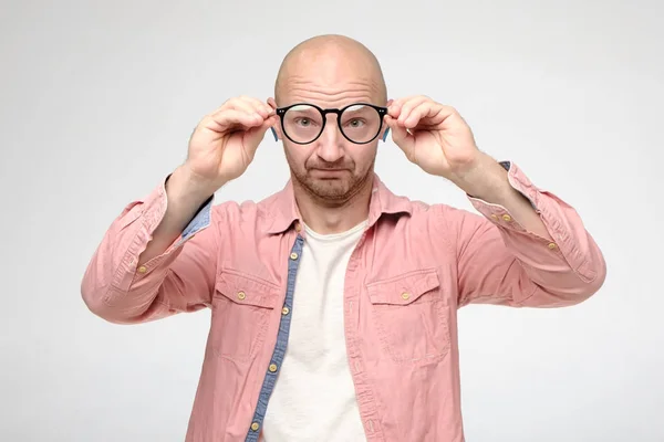 Bald, unshaven man took off glasses, keeping them at a distance, he examines the cleanliness of the lenses and looks funny. — Stock Photo, Image