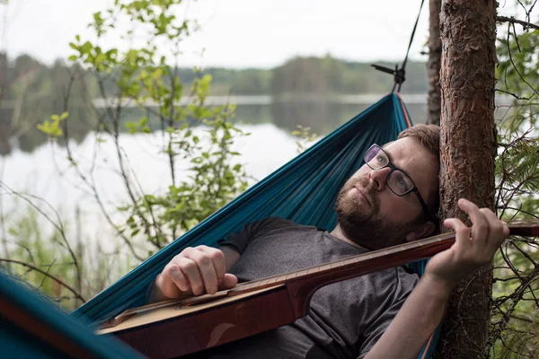 Triste hombre yace en una hamaca con una guitarra acústica en las manos, toca una melodía y mira soñadoramente al cielo, en la orilla de un lago . — Foto de Stock