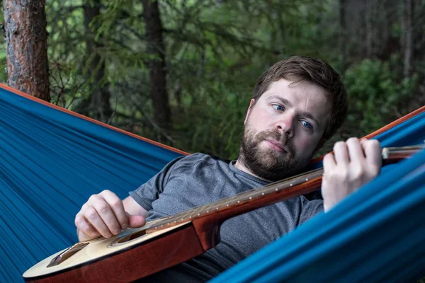 El hombre barbudo pensativo miente en una hamaca con una guitarra acústica en las manos y toca una melodía contra un bosque . — Foto de Stock