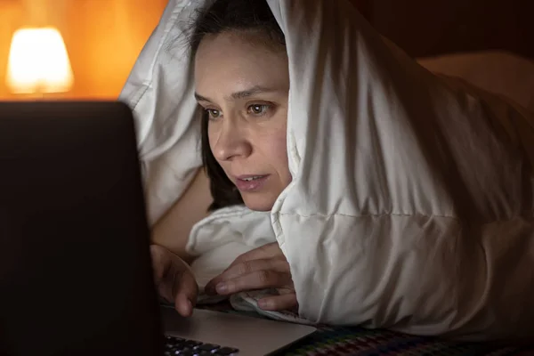 Woman looks with interest at a laptop, hiding under a blanket, in bed, at night by the light of a lamp. — Stockfoto