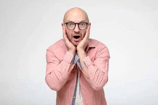 Geschokte kale man met bril houdt emotioneel handen op zijn gezicht, opent in verrassing zijn mond en kijkt met grote ogen in de camera. — Stockfoto