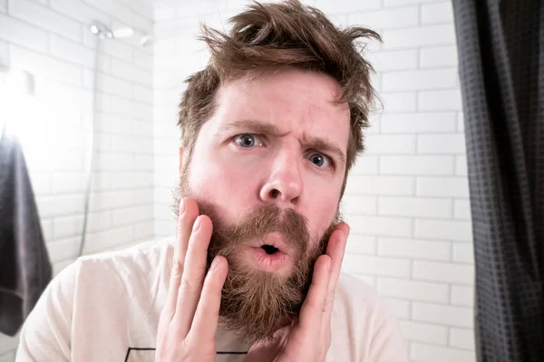 Overgrown man, with a shaggy hairstyle and beard, sees himself reflected in the mirror, he is shocked by his appearance. — Stock Photo, Image