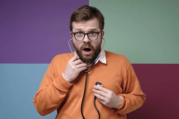 Excited bearded man listens to his heart with a stethoscope, he opened mouth in amazement and looks with fear at the camera.
