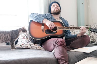 Hayalperest adam, gözleri kapalı, gitar çalıyor, yumuşak bir kanepede oturuyor, yanında neler olduğuyla ilgilenen bir kedi var..