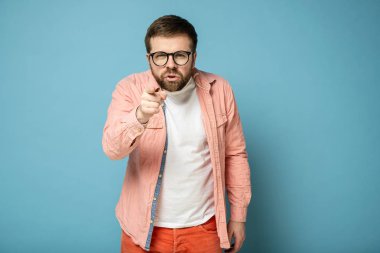 Canı sıkkın beyaz bir adam işaret parmağıyla birini işaret ediyor ve kameraya gözlerini kısarak bakıyor..
