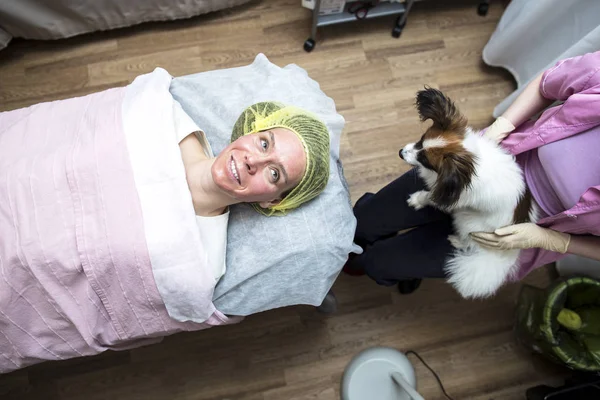 Kobieta na wizycie kosmetyczki robi odmładzający zabieg twarzy, peeling chemiczny i spojrzenia, uśmiech, do jej zmartwionego psa. — Zdjęcie stockowe