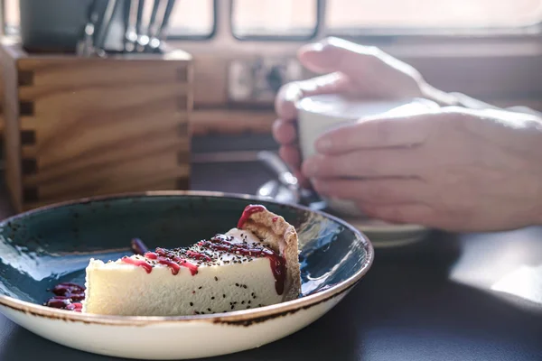 Νόστιμο cheesecake σε ένα πιάτο, με φόντο τα χέρια κρατώντας ένα φλιτζάνι ευωδιαστό καπουτσίνο, υπό φυσικό φως από. — Φωτογραφία Αρχείου