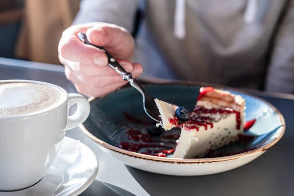 Cheesecake με φρέσκο μούρο σε ένα κουτάλι, με φόντο ένα κομμάτι σε ένα πιάτο, δίπλα σε ένα φλιτζάνι καπουτσίνο, σε φυσικό φως. — Φωτογραφία Αρχείου