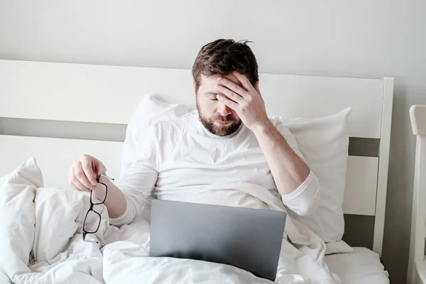 Mannen i pyjamas i sängen med en laptop, tog av sig glasögon, blundade och höll hand på pannan, arbetade hela natten och var trött. — Stockfoto