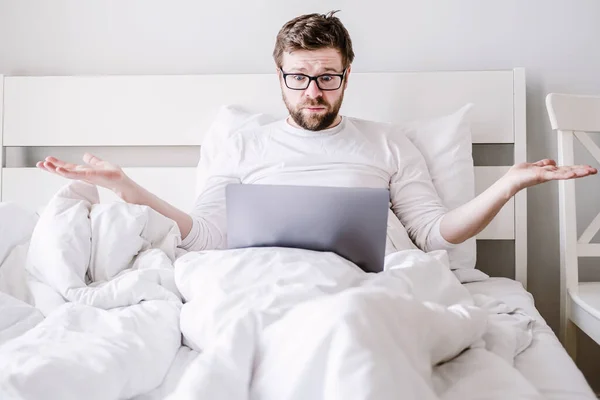 Upprörd man i pyjamas i sängen med en laptop, stirrar han på skärmen i förvirring och gör händerna åt sidorna. — Stockfoto