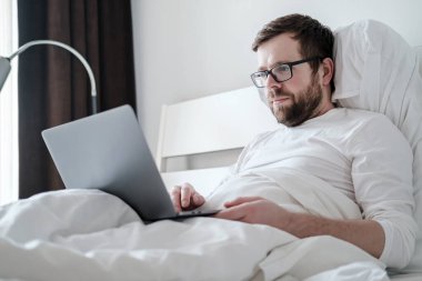 Freelancer sabahı. Tatmin olmuş, pijamalı genç bir adam laptopla çalışıyor, yumuşak bir yatakta, rahat dairelerde yatıyor..