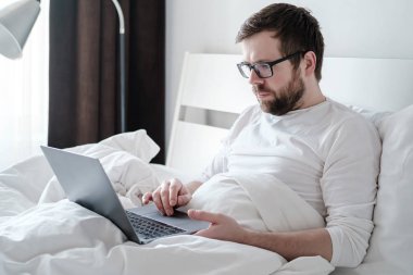 Freelancer sabahı. Pijamalı odaklanmış bir adam laptopla çalışır, yumuşak bir yatakta, rahat dairelerde, doğal ışıkta yatar..