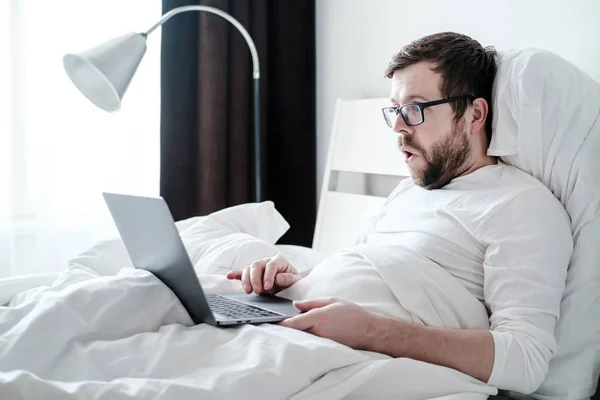 Morgon av en framgångsrik frilansare. Överraskad, nöjd man i pyjamas arbetar med en bärbar dator, ligger i en mjuk säng, i naturligt ljus — Stockfoto