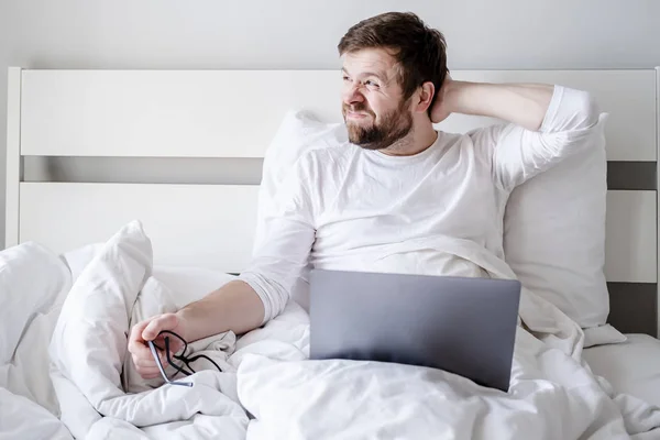 Творческий процесс. Блогер отвлекается от работы с ноутбуком, снимает очки и задумчиво смотрит в окно в постели . — стоковое фото